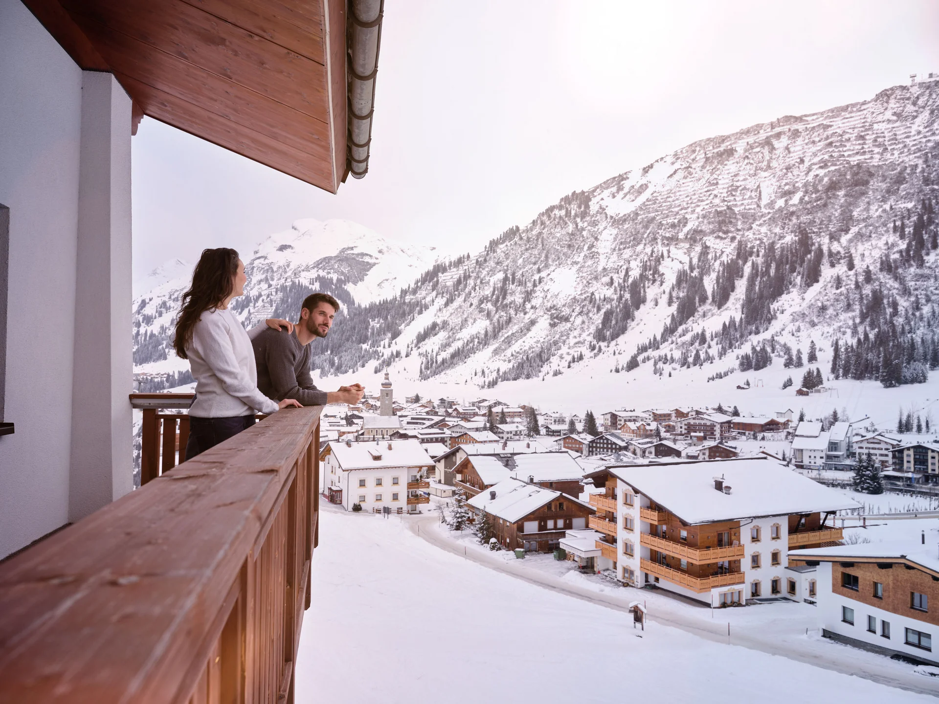 Zwei Personen stehen auf einem Balkon mit Blick auf verschneites Dorf und Berge