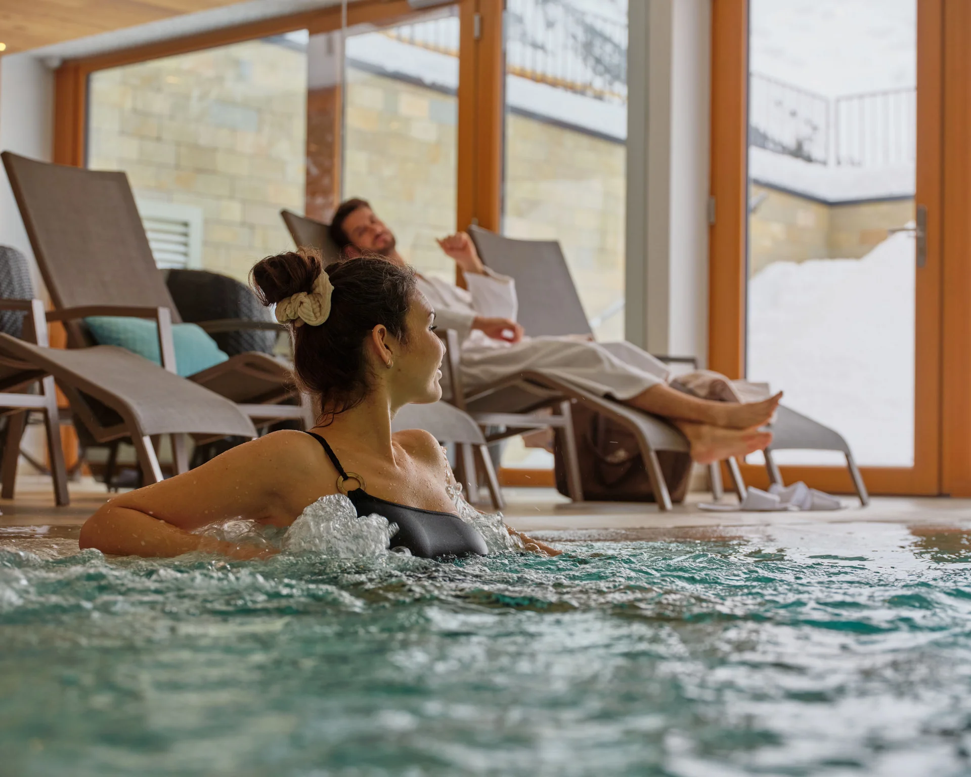 Frau entspannt im Whirlpool, Mann liegt auf Liegestuhl im Innenbereich mit großen Fenstern