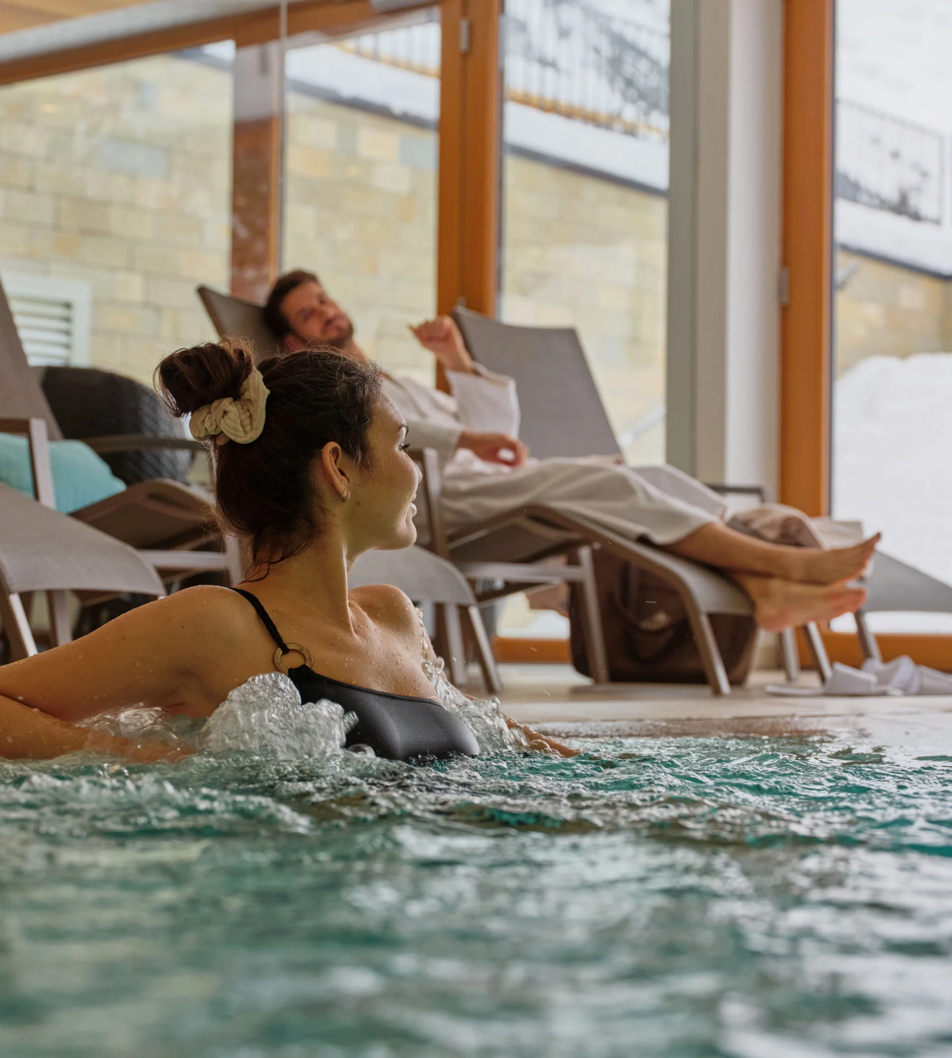 Frau entspannt im Whirlpool, Mann liegt auf Liegestuhl im Innenbereich mit großen Fenstern