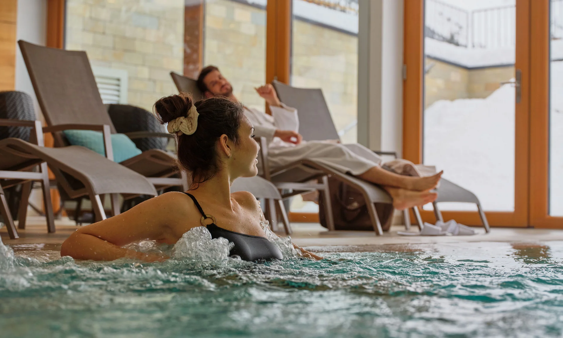 Frau entspannt im Whirlpool, Mann liegt auf Liegestuhl im Innenbereich mit großen Fenstern