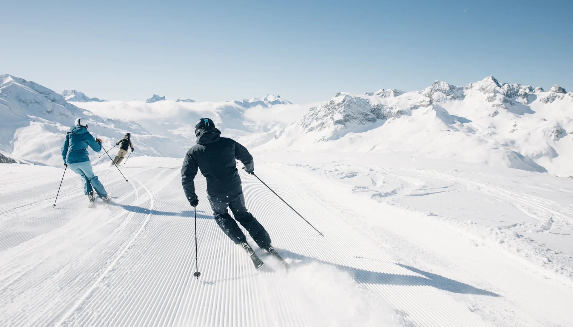 Skifahrer fahren auf präparierter Piste in verschneiter Berglandschaft bei klarem Himmel