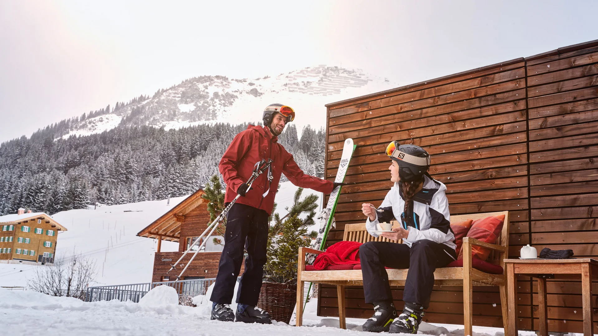 Zwei Skifahrer im Winter vor einer Hütte mit schneebedeckten Bergen im Hintergrund