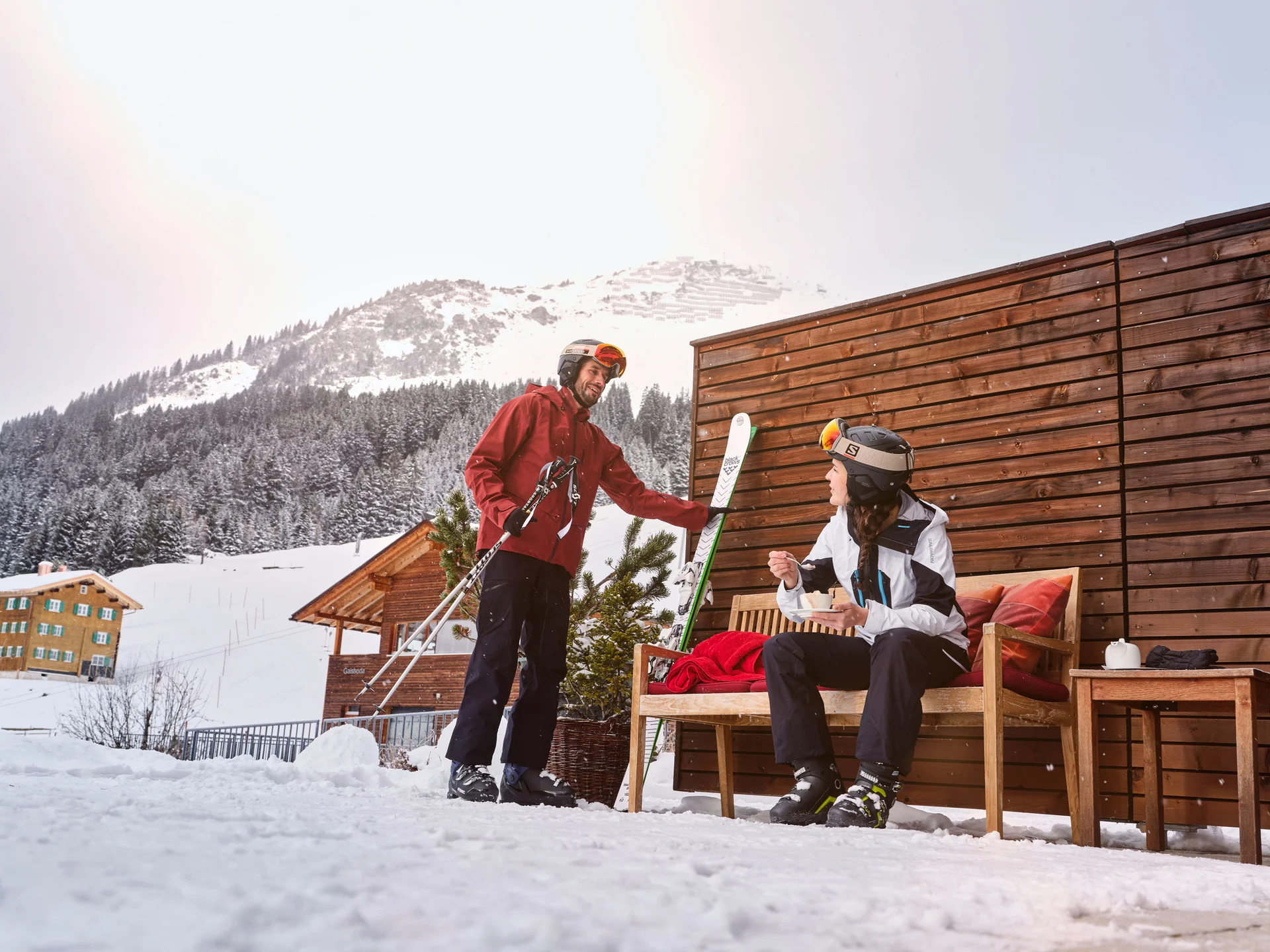 Zwei Skifahrer im Winter vor einer Hütte mit schneebedeckten Bergen im Hintergrund