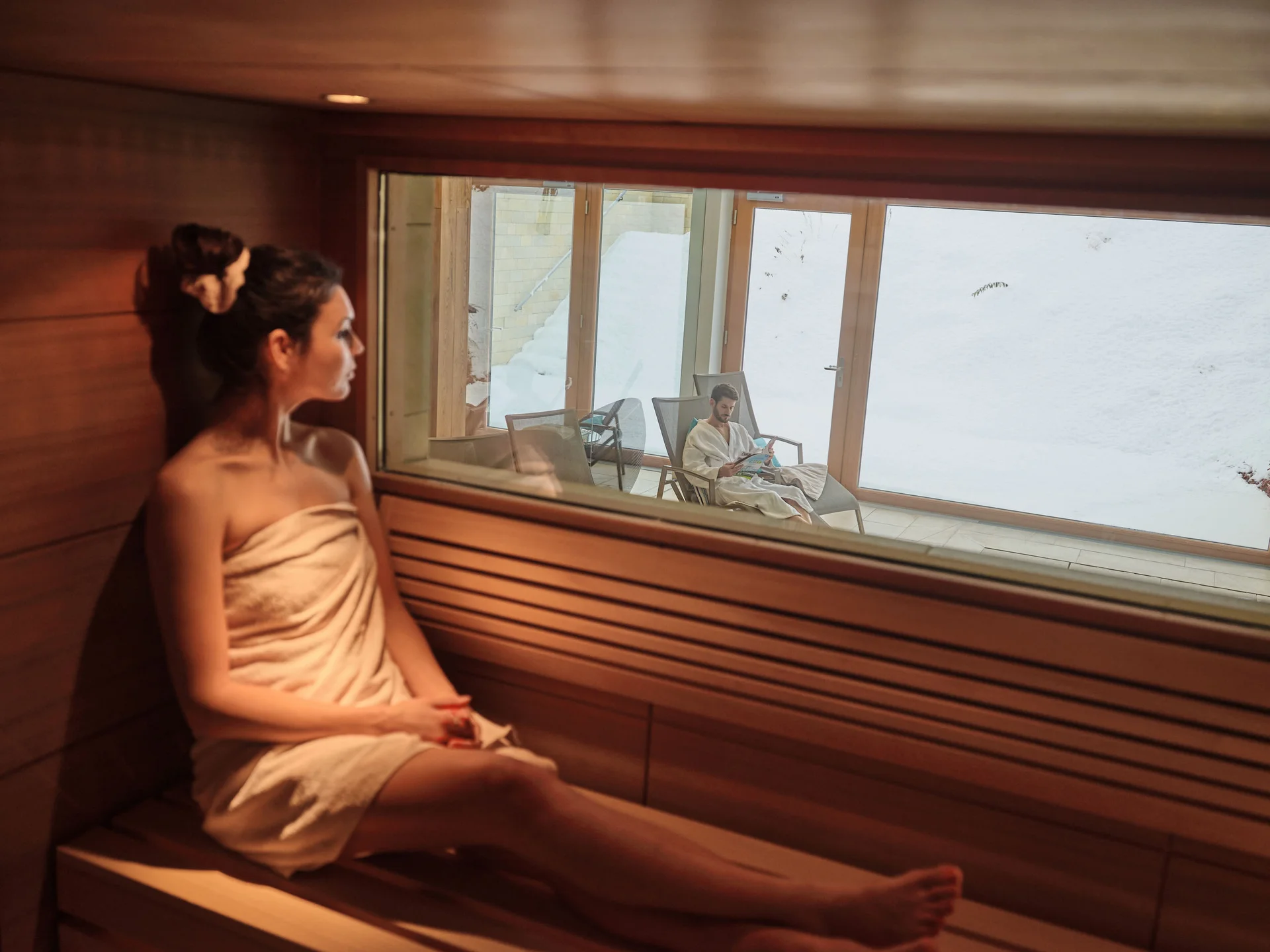 Frau in Sauna blickt durch Fenster auf Mann in Bademantel draußen im Schnee