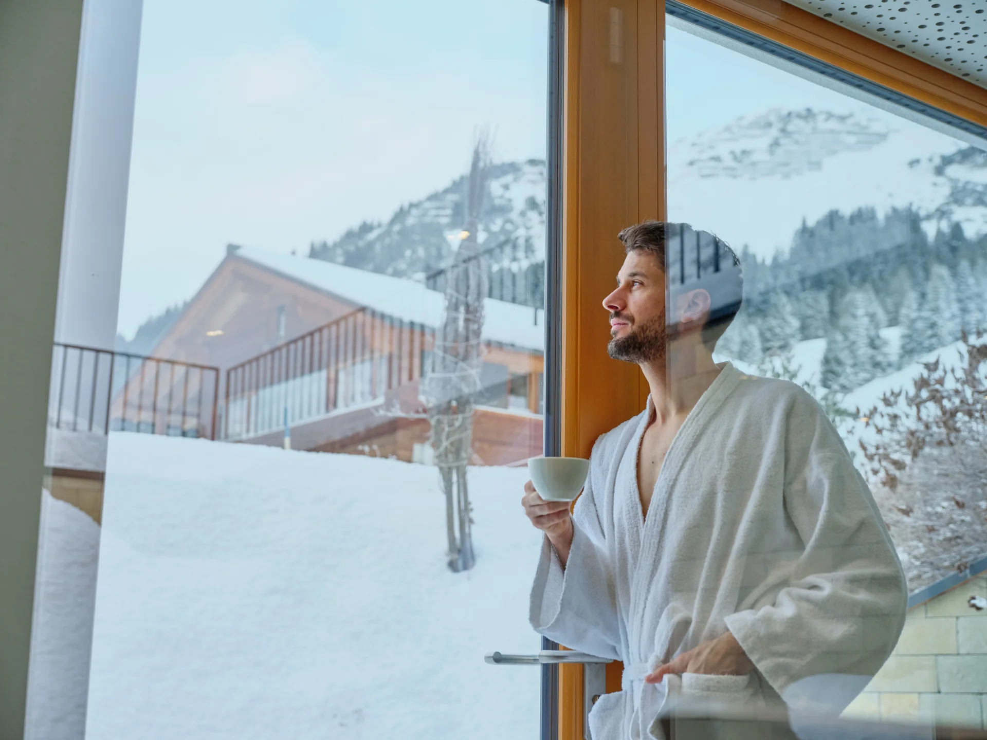 Bilder und Impressionen aus dem Sunora Mann im Bademantel trinkt Kaffee und schaut aus dem Fenster auf Schneeberge