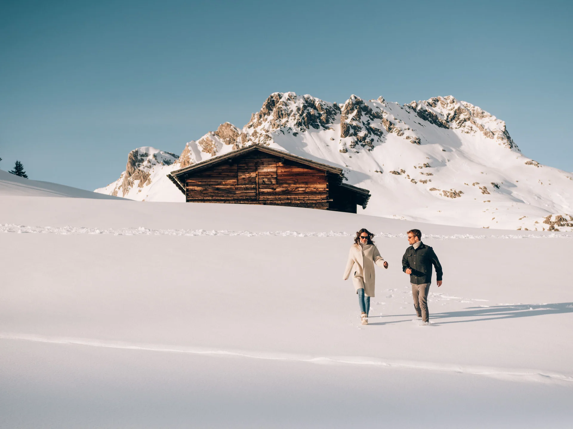 Paar spaziert im Schnee vor einer Holzhütte mit verschneiten Bergen im Hintergrund