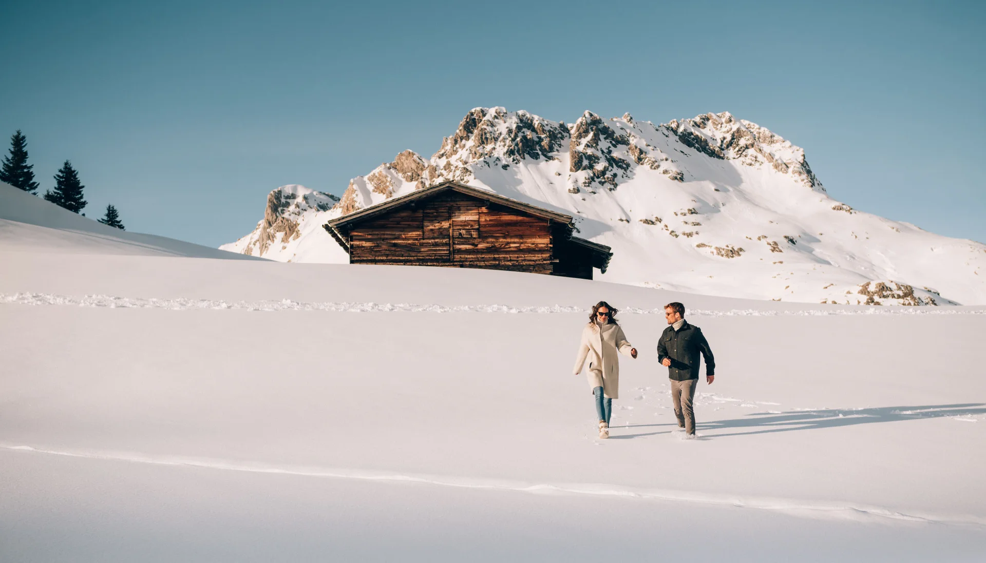 Paar spaziert im Schnee vor einer Holzhütte mit verschneiten Bergen im Hintergrund