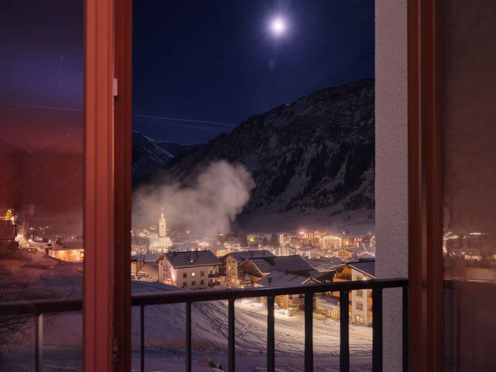 Bilder und Impressionen aus dem Sunora Blick durch Fenster auf verschneites Dorf und Berge bei Nacht mit Vollmond