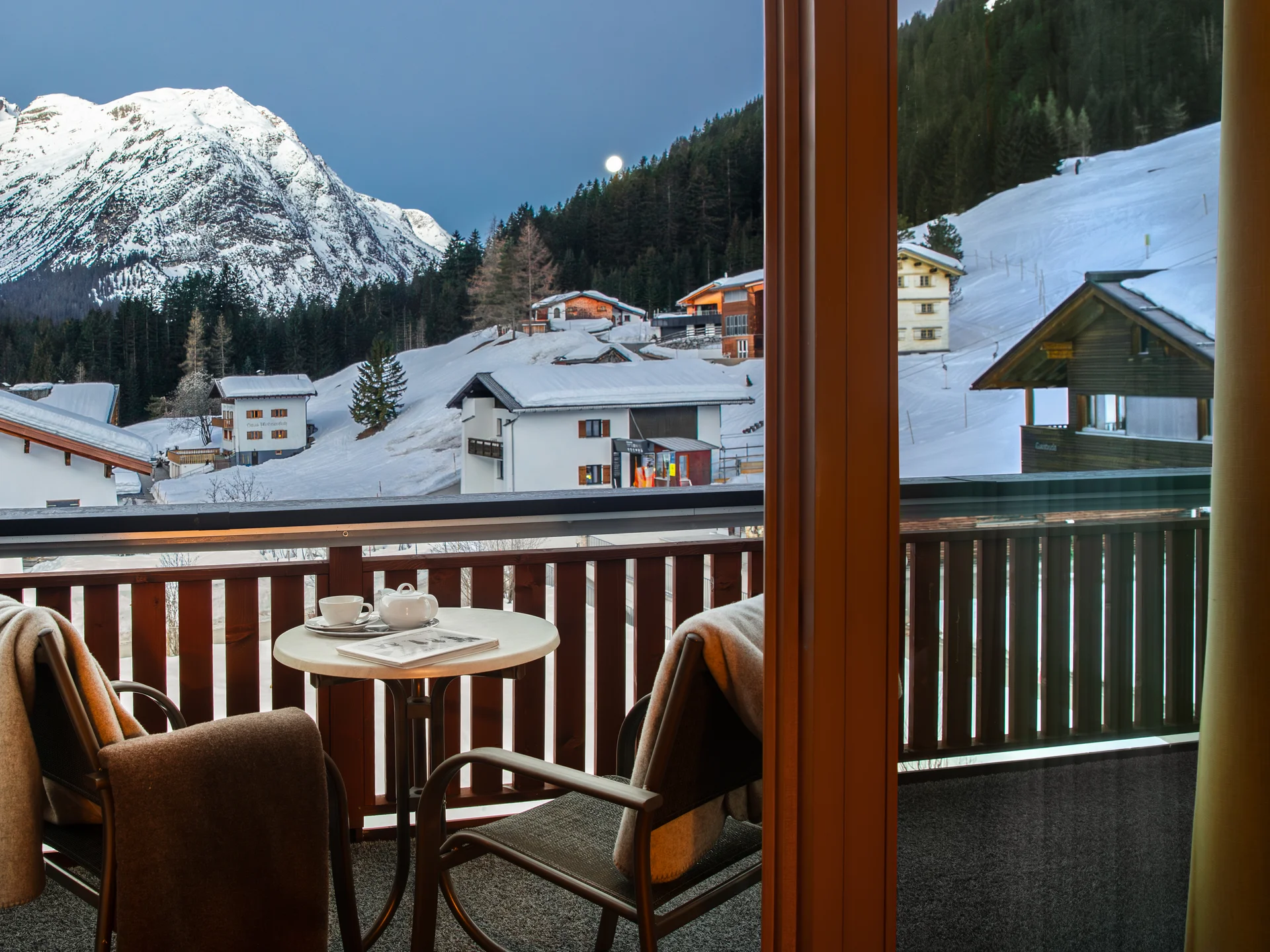 Bilder und Impressionen aus dem Sunora Balkon mit Tisch und Stühlen mit Blick auf schneebedeckte Berge und Häuser