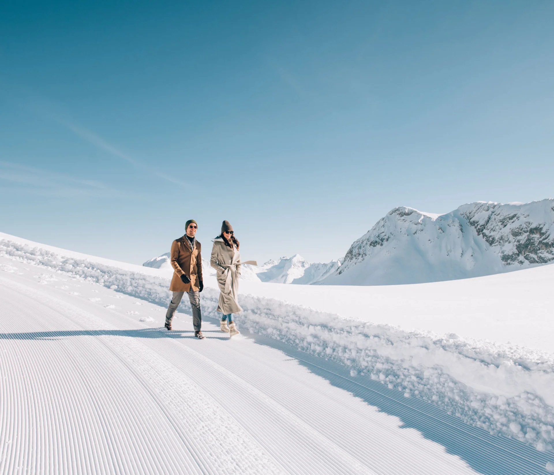 Zwei Menschen gehen auf einem verschneiten Weg mit Bergpanorama bei klarem Himmel spazieren