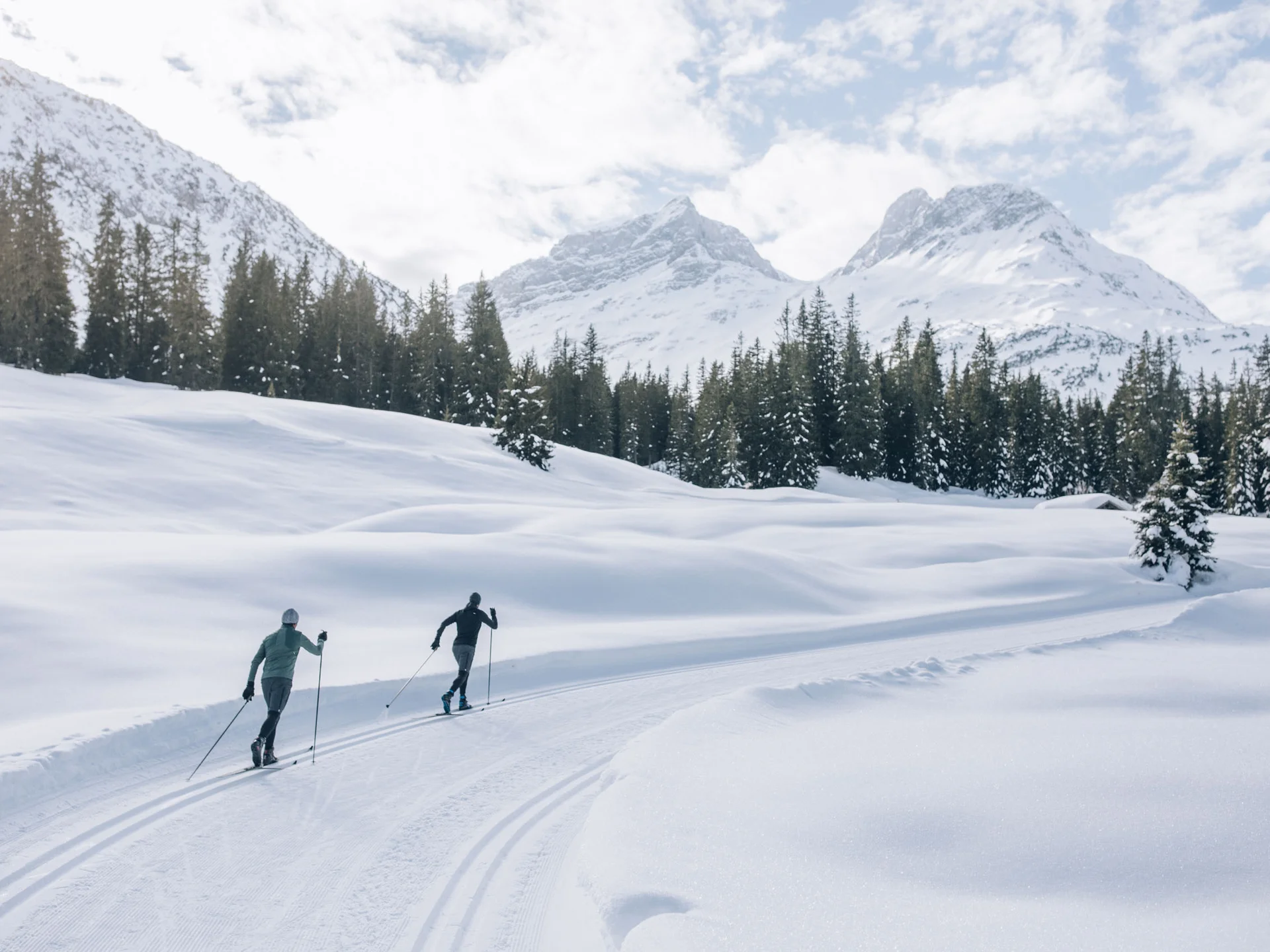 Zwei Skifahrer in verschneiter Berglandschaft mit Tannen und bewölktem Himmel